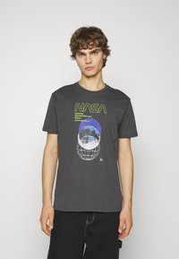 T-shirt en coton gris avec un graphique de la NASA comprenant du texte, des designs de globe et de lune en bleu et blanc. Manches courtes et col rond.