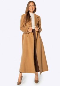 Dlouhý kabát v barvě velbloudí z vlněné směsi s dvojitým zapínáním, klopovými límci a hlubokými předními kapsami. Stylizovaný přes krémový svetr a hnědé kalhoty.