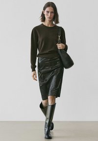 Brauner Pullover, glänzender schwarzer Bleistiftrock, kniehohe schwarze Stiefel und eine schwarze Ledertasche. Glatte Texturen und figurbetonte Silhouette in einem minimalistischen Ambiente.