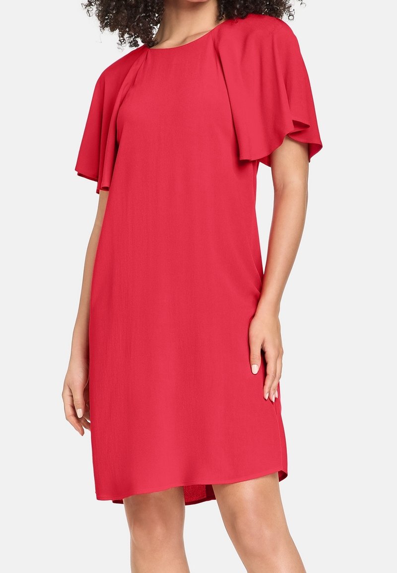 Femme portant une robe rouge vif mi-longue avec des manches larges et aériennes et un col rond, posant devant un fond blanc uni.