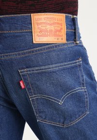 Jeans de mezclilla azul oscuro con un parche de cuero, costuras de cobre y una pequeña etiqueta roja en el bolsillo trasero. Estilo 511, tamaño W32 L32.