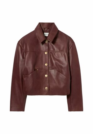 Femme debout les mains dans les poches, portant une veste en cuir marron, un col roulé rouge, une longue jupe à motifs et des bottes noires sur un fond uni.