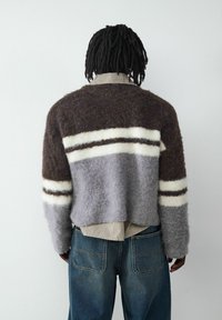 Gestreifter Wollpullover in Brauntönen, Weiß und Grau mit einer flauschigen Textur. Über einem karierten Hemd getragen, kombiniert mit Jeanshosen.