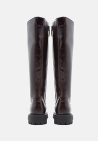 Um par de botas altas de couro castanho escuro com solas pretas robustas, fechos éclair visíveis na parte de trás e um logótipo discreto perto do calcanhar.