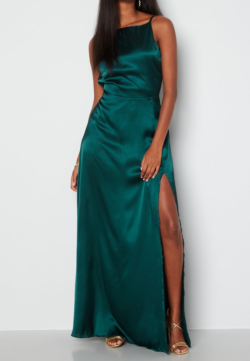 Femme portant une robe de soirée en satin vert émeraude sans manches, fendue jusqu'à la cuisse, avec des talons dorés à lanières, debout sur un fond blanc.