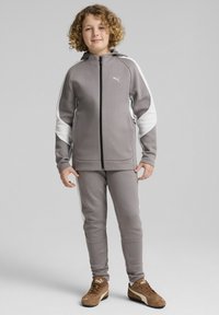 Grå atletisk træningsdragt med hvide side striber, bestående af en jakke med lynlås og matchende bukser. Et par brune sneakers fuldender looket.
