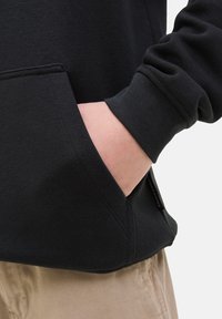 Hoodie noir en tissu texturé avec une poche kangourou et des poignets côtelés, associé à un pantalon beige clair.
