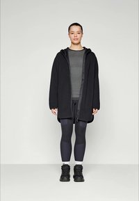 Zwarte hooded jasje met een zachte textuur, gedragen over een donkergrijze aansluitende top en zwarte leggings. Schoeisel zijn zwarte sportschoenen met een robuuste zool.