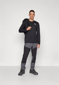 Svart langermet skjorte, grå og svarte bukser med detaljpaneler, svart fottøy og en svart ryggsekk. Nøytral bakgrunn.