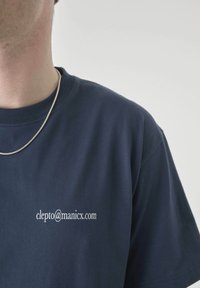 Person wearing a navy blue T-shirt with white email text "clepto@manicx.com" and a thin silver chain necklace, shown from shoulders up.