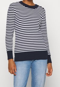 Pull en maille rayé bleu marine et blanc avec ourlet et poignets côtelés. Détails de boutons sur une épaule. Associé à un jean bleu clair.