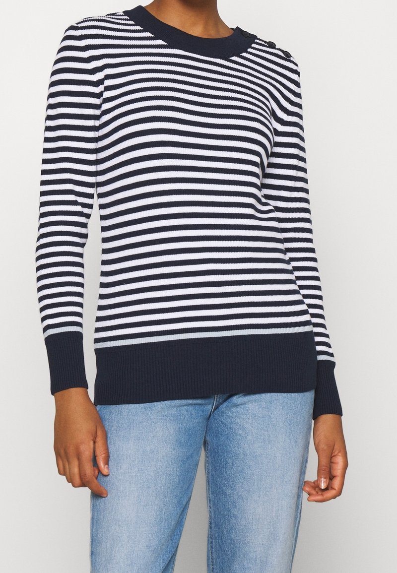Pull en maille rayé bleu marine et blanc avec ourlet et poignets côtelés. Détails de boutons sur une épaule. Associé à un jean bleu clair.