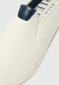 Fehér perforált anyagból készült slip-on sneaker, tengerészkék bőr díszítéssel a saroknál. Gumi talppal és varrott részletekkel rendelkezik.