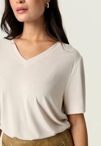 Vaaleanbeige v-kaula-aukkoinen t-paita, joka on valmistettu pehmeästä kankaasta. Paidassa on lyhyet hihat ja rento istuvuus. Tasainen pinta jossa on hienovaraisia ompeluyksityiskohtia.