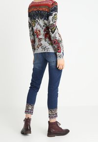 Pull en tricot avec imprimé floral, gris avec des accents rouges, bleus et verts ; jeans bleus avec des poignets à motifs ; bottines bordeaux.