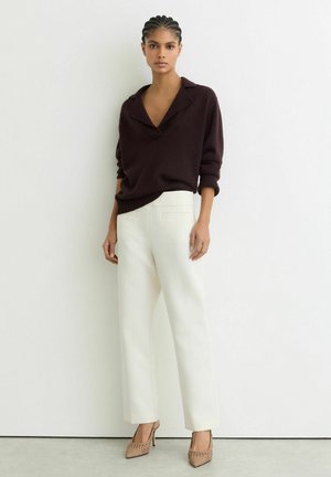 Fonott hajú nő sötét hosszú ujjú pulóverben, fehér magas derekú nadrágban és bézs, sarokkal ellátott, szőtt cipőben áll fehér fal előtt.