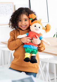 Kind met krullend haar in een bruine hoodie houdt een knuffelpop vast. De pop heeft oranje staarten, een rode top, groene broek en zwarte schoenen.