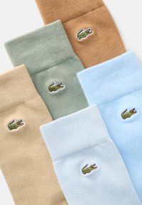 Cinco pares de calcetines en beige, verde, marrón y azul claro, con un pequeño logo de cocodrilo bordado en cada uno. Están hechos de material suave.