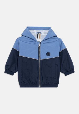 Veste coupe-vent bleu et marine avec capuche, fermeture éclair à l'avant, poignets élastiques et deux poches latérales. Elle présente un logo sur la poitrine et des coutures contrastantes.