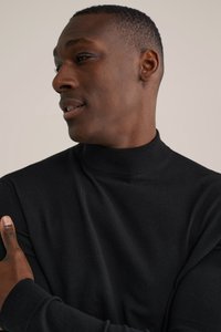 Melns ribots augstas apkakles džemperis ar piegulošu dizainu, maigu tekstūru un garām piedurknēm, ko valkā persona ar īsiem, nogrieztiem matiem.