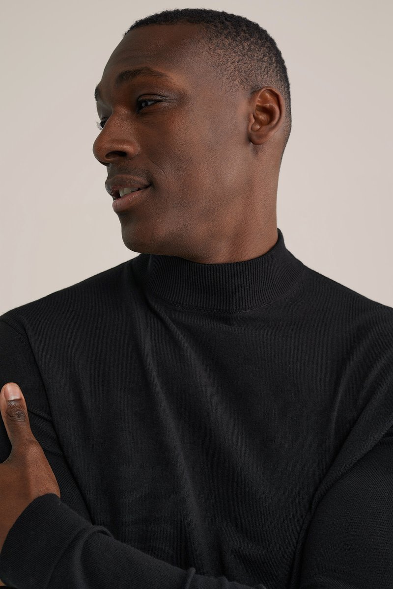 Melns ribots augstas apkakles džemperis ar piegulošu dizainu, maigu tekstūru un garām piedurknēm, ko valkā persona ar īsiem, nogrieztiem matiem.