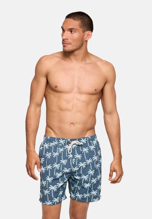 Hombre sin camiseta con pelo corto, vistiendo pantalones cortos de baño azules con patrón de palmeras blancas, de pie sobre un fondo sencillo y claro, mirando hacia la izquierda.