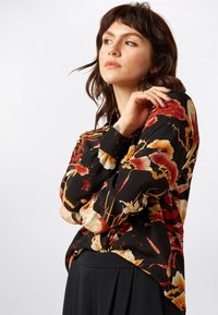 Langarmbluse aus schwarzem Stoff mit lebhaften floralen Mustern in Rot und Creme, mit lockerer Passform und Knopfmanschetten.