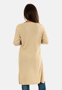 Donna con lunghi capelli castani che indossa un cardigan lungo beige a costine e jeans blu, girata di spalle su uno sfondo chiaro e uniforme.