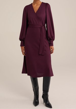 Abito avvolgente color malva con maniche lunghe a sbuffo, tessuto strutturato e cintura annodata in vita. Abbinato a stivali neri al ginocchio e taglio aderente.