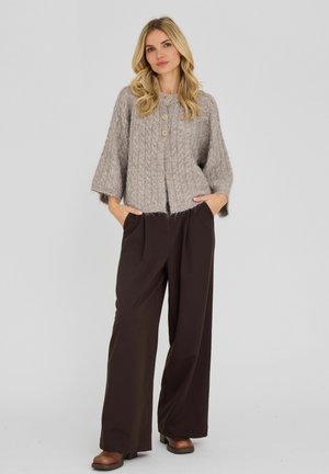 Femme aux longs cheveux blonds portant un cardigan texturé gris avec des boutons, un pantalon large marron foncé et des chaussures marron, debout, les mains dans les poches.