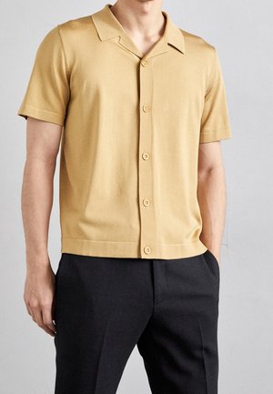 Koszula z krótkim rękawem i kołnierzykiem, wykonana z miękkiego, beżowego materiału o gładkiej teksturze i z zapięciem na guziki, zestawiona z ciemnymi spodniami.