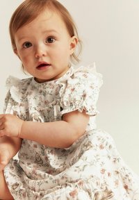 Weißes Kleid mit floralen Mustern und Hasenmotiven, mit rüschigen Ärmeln und Saum, getragen von einem sitzenden Kind mit hellbraunen Haaren.