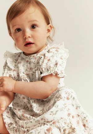 Weißes Kleid mit floralen Mustern und Hasenmotiven, mit rüschigen Ärmeln und Saum, getragen von einem sitzenden Kind mit hellbraunen Haaren.