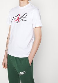 Person som bär en vit T-shirt med "Flight" och en röd basketlogo, ihop med gröna Nike sweatpants, med handen i fickan.