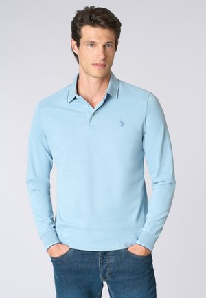 Uomo con capelli scuri che indossa una polo a maniche lunghe blu chiaro e jeans blu, in piedi con le mani nelle tasche su uno sfondo semplice.