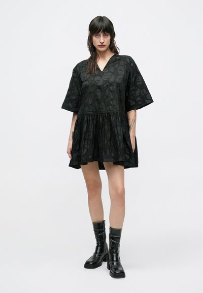 Μαύρο φόρεμα με κοντά μανίκια και V-λαιμόκοψη, υφασμένο με υφή που απεικονίζει σχέδιο φύλλων και φουσκωτή φούστα. Φορεμένο με μαύρες μπότες μέχρι το γόνατο.