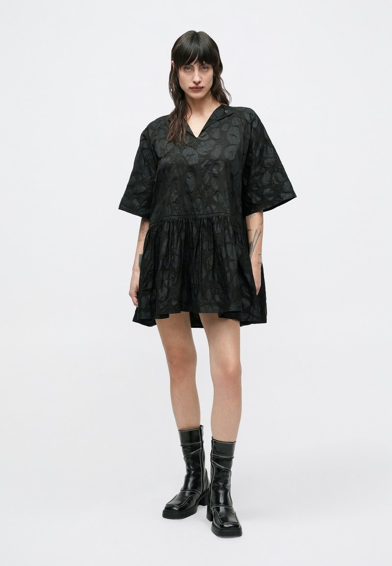 Robe noire à manches courtes avec un col en V, tissu texturé présentant un motif de feuilles et une jupe évasée. Portée avec des bottes noires hautes jusqu'aux genoux.