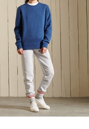 Blau strukturierter Pullover mit Rundhalsausschnitt, kombiniert mit grauen Jogginghosen mit rot-weiß gestreiften Bündchen und weißen Sneakers.