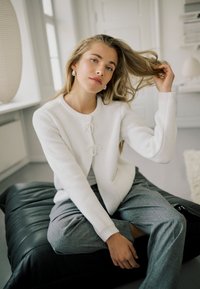 Cardigan blanc texturé avec fermetures à nouer semblables à des perles, associé à un pantalon gris, assis sur un coussin noir, dans une pièce lumineuse.