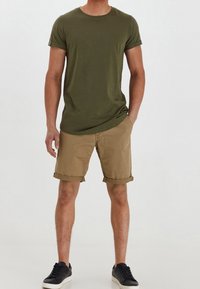 Uomo che indossa una maglietta a maniche corte verde oliva, pantaloni corti khaki arrotolati e sneaker nere con suole bianche, in piedi con una mano in tasca.