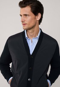 Cárdigan negro con diseño de escote en V, botonadura frontal y puños acanalados. Lleva puesto sobre una camisa de rayas azul claro, con una textura de tela suave.