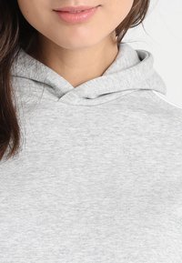 Jonge vrouw met lang bruin haar, gekleed in een lichtgrijze capuchontrui, lippen licht geopend in een neutrale uitdrukking.