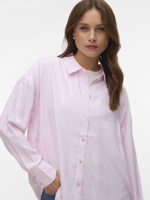 Młoda kobieta o brązowych włosach nosi luźną, różowo-białą, paskowaną koszulę zapinaną na guziki i ma jedną rękę w kieszeni.