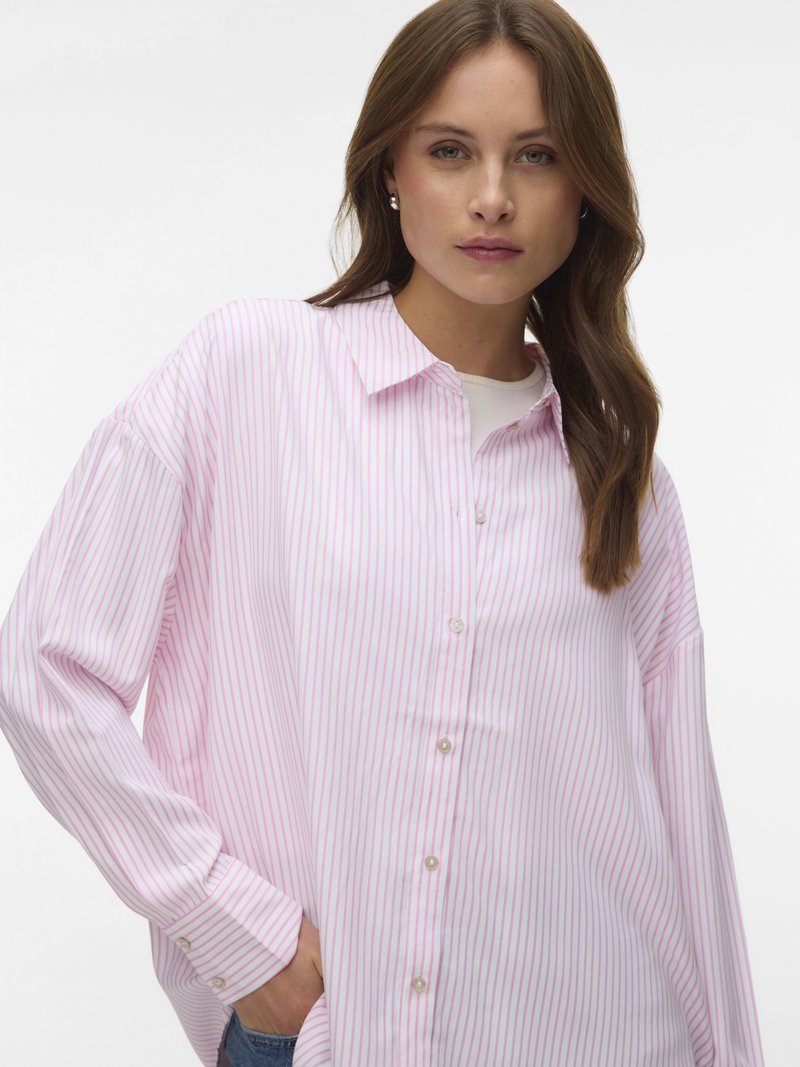Mladá žena s hnědými vlasy nosí volnou růžovo-bílou proužkovanou košili na knoflíky a jednu ruku má v kapse.