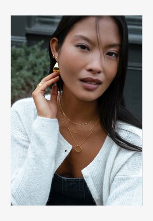 Kuldsed ehted, mis hõlmavad kihilisi kaelakette ümmarguse ripatsiga, rõngaskõrvarõngaid ja kudumitest valget kardigaani. Siledad tekstuurid ja stiilne disain.