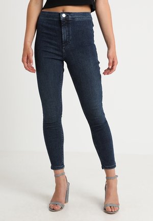 Woman wearing dark blue skinny jeans and silver high-heeled ankle-strap sandals, standing against plain light background.