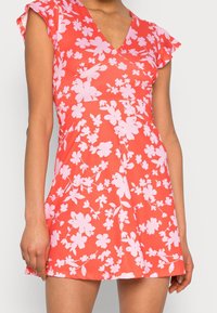 Robe florale avec un décolleté en V, manches courtes volantes, en corail avec des fleurs roses. Tissu lisse, corsage ajusté, jupe évasée.