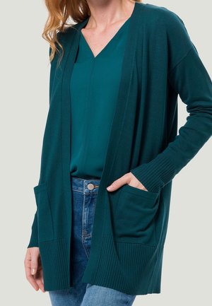 Frau trägt eine türkisfarbene, offen geschnittene Strickjacke über einer türkisfarbenen Bluse und blauen Jeans, mit der rechten Hand in der Tasche der Strickjacke.