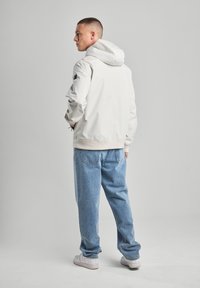 Giacca con cappuccio grigia chiara con polsini ed orlo elastici, abbinata a jeans denim blu ampia e sneaker bianche. Vista posteriore che mostra un design minimalista.