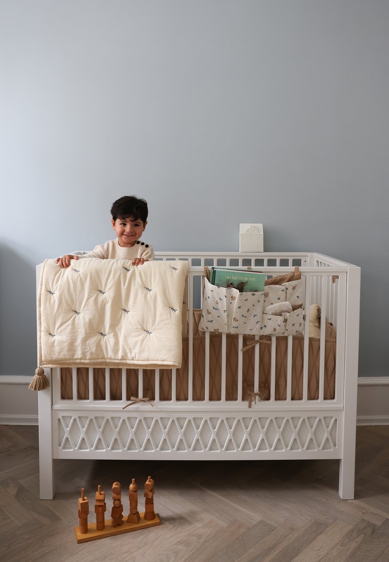 Νήπιο σε λευκό κούνια κρατώντας μπεζ κουβέρτα, με υφασμάτινο οργανωτή και βιβλία μέσα, ξύλινο παιχνίδι στοιβαγής στο πάτωμα σε δωμάτιο βρεφονηπιακού σταθμού.